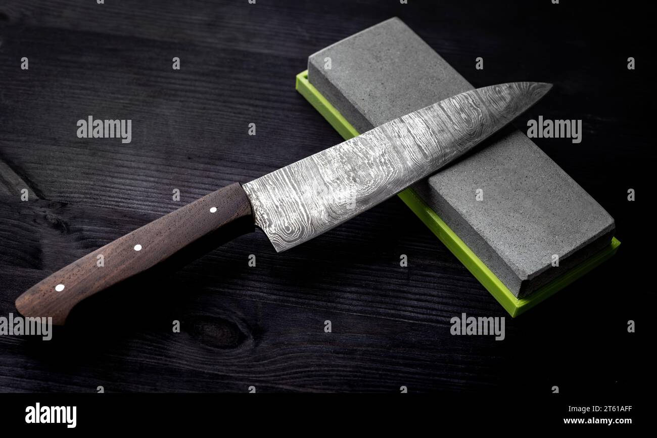 Il coltello da cucina Big si appoggia sulla pietra affilante sul tavolo da cucina sullo sfondo nero Foto Stock