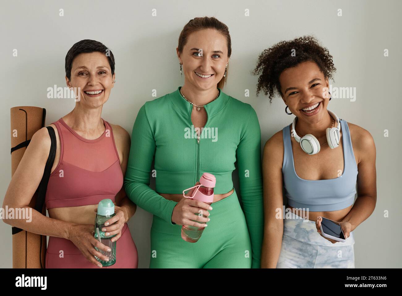 Ritratti in vita di tre donne spensierate che sorridono alla macchina fotografica in palestra e tengono in mano bottiglie d'acqua Foto Stock
