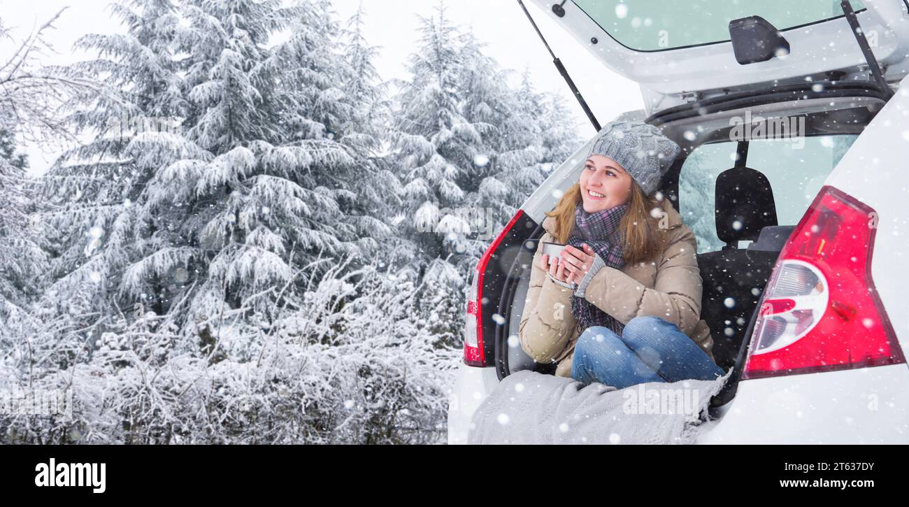 Una donna con un cappello di lana siede nel bagagliaio dell'auto e tiene una tazza di tè caldo in mano. Foto Stock