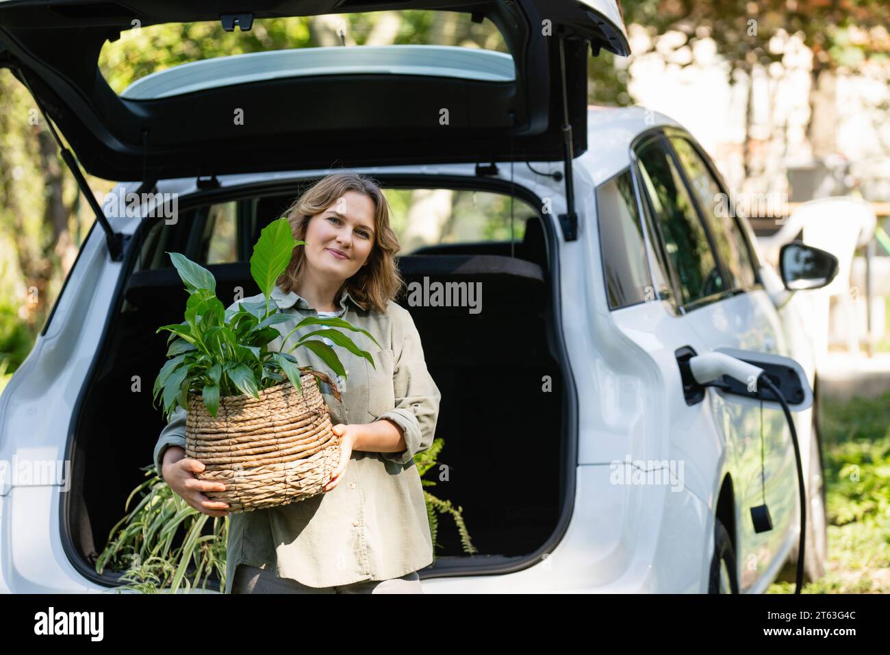 Donna con impianto in vaso vicino a un'auto elettrica di ricarica nel cortile di una casa di campagna. Foto Stock