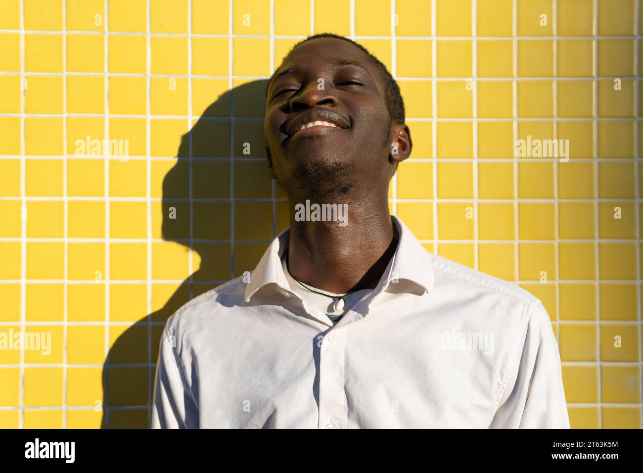 Allegro giovane uomo nero che ride contro un muro piastrellato giallo vibrante, con il sole che gli proietta un'ombra Foto Stock