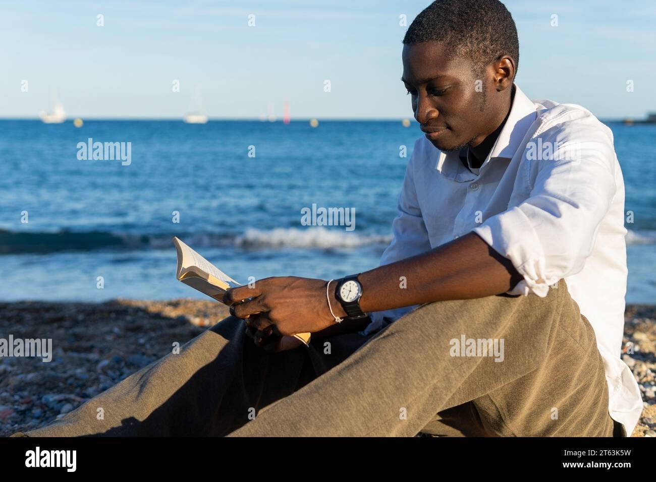 Vista laterale di un giovane afroamericano immerso in un libro seduto sulla spiaggia con onde dolci e barche a vela distanti sullo sfondo Foto Stock
