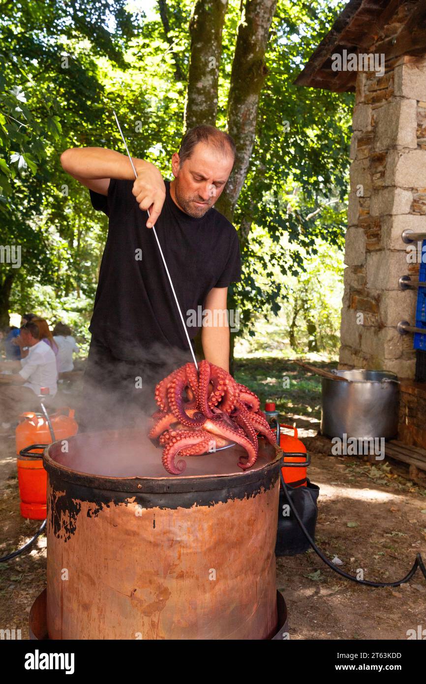 Lo chef attento in abiti scuri solleva un polpo appena bollito da una pentola rustica, mandando pennacchi di vapore in aria contro l'ambiente con la schiena sfocata Foto Stock