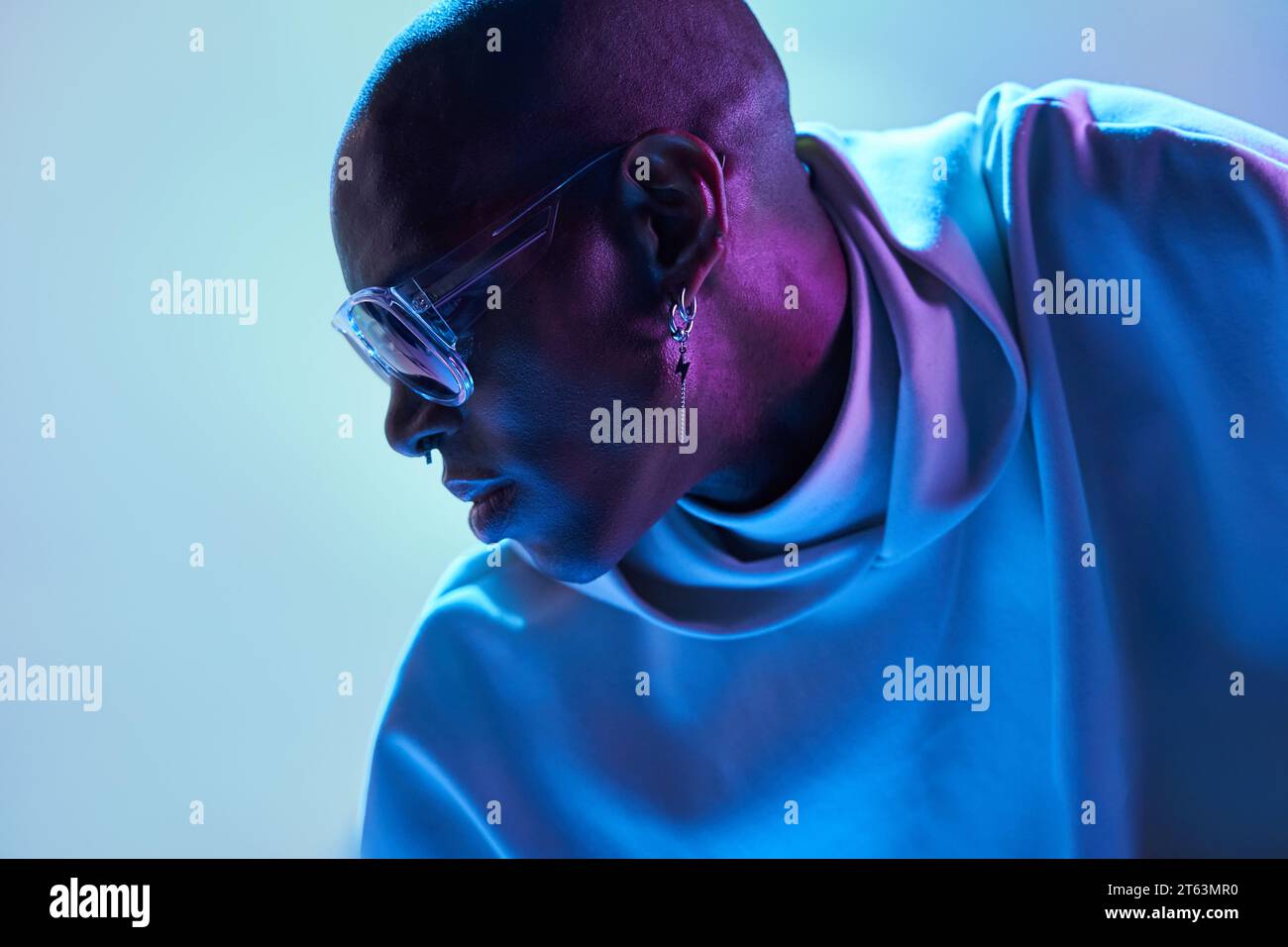 Uomo calvo afro-americano che indossa orecchini e occhiali da sole e guarda in basso in uno studio moderno con luci illuminanti Foto Stock