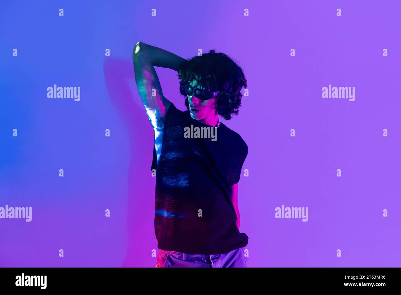 Un giovane latino con i capelli ricci mette in posa dinamica su uno sfondo viola vibrante, illuminato da luci al neon che riflettono il suo abbigliamento Foto Stock