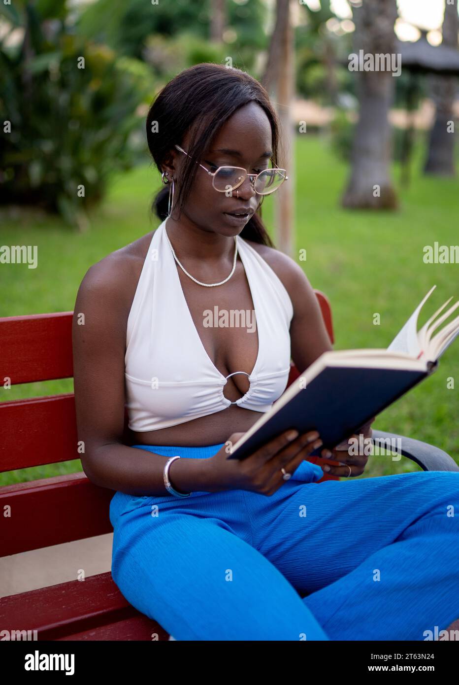 Donna nera travolgente adornata con top bianco e pantaloni blu che si concentrano profondamente sul suo libro mentre si siede su una panchina rossa in un lussureggiante giardino verde Foto Stock