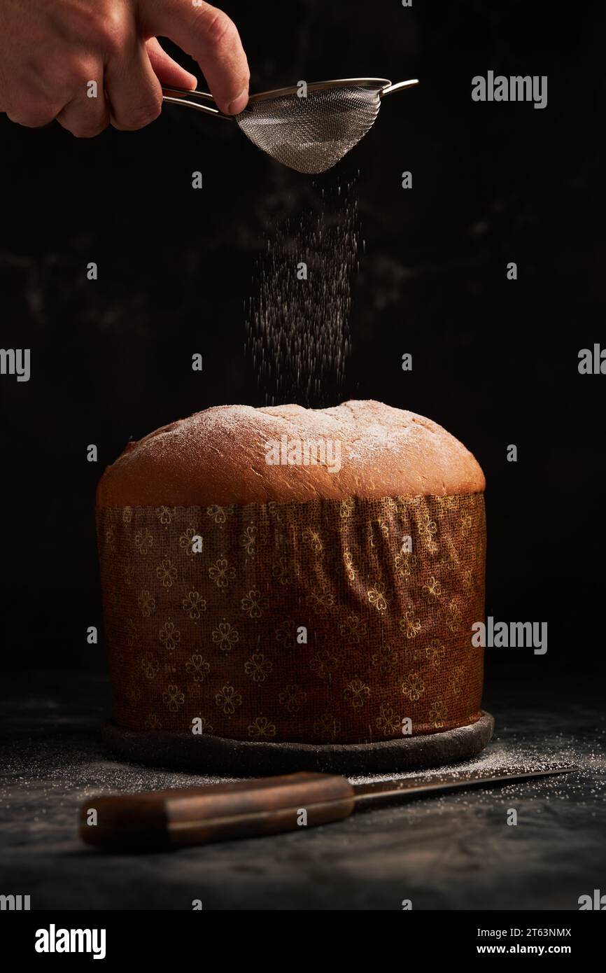 Una mano spolvera delicatamente lo zucchero in polvere su un panettone, catturando la neve a mezz'aria in un ambiente buio Foto Stock