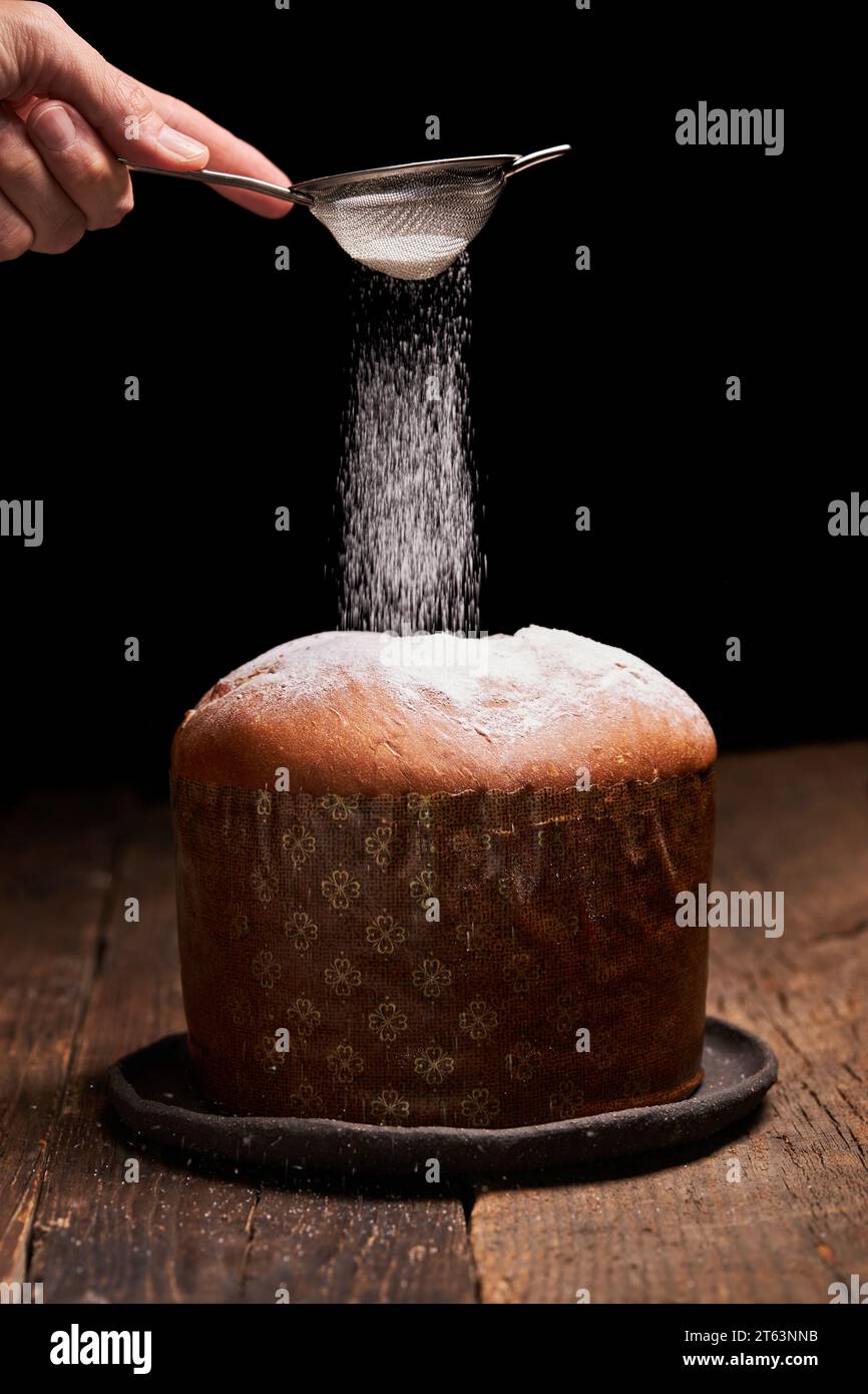 Una mano setacciava con grazia lo zucchero a velo sopra un classico panettone, enfatizzando la delicata consistenza del dessert e l'essenza festosa. Foto Stock