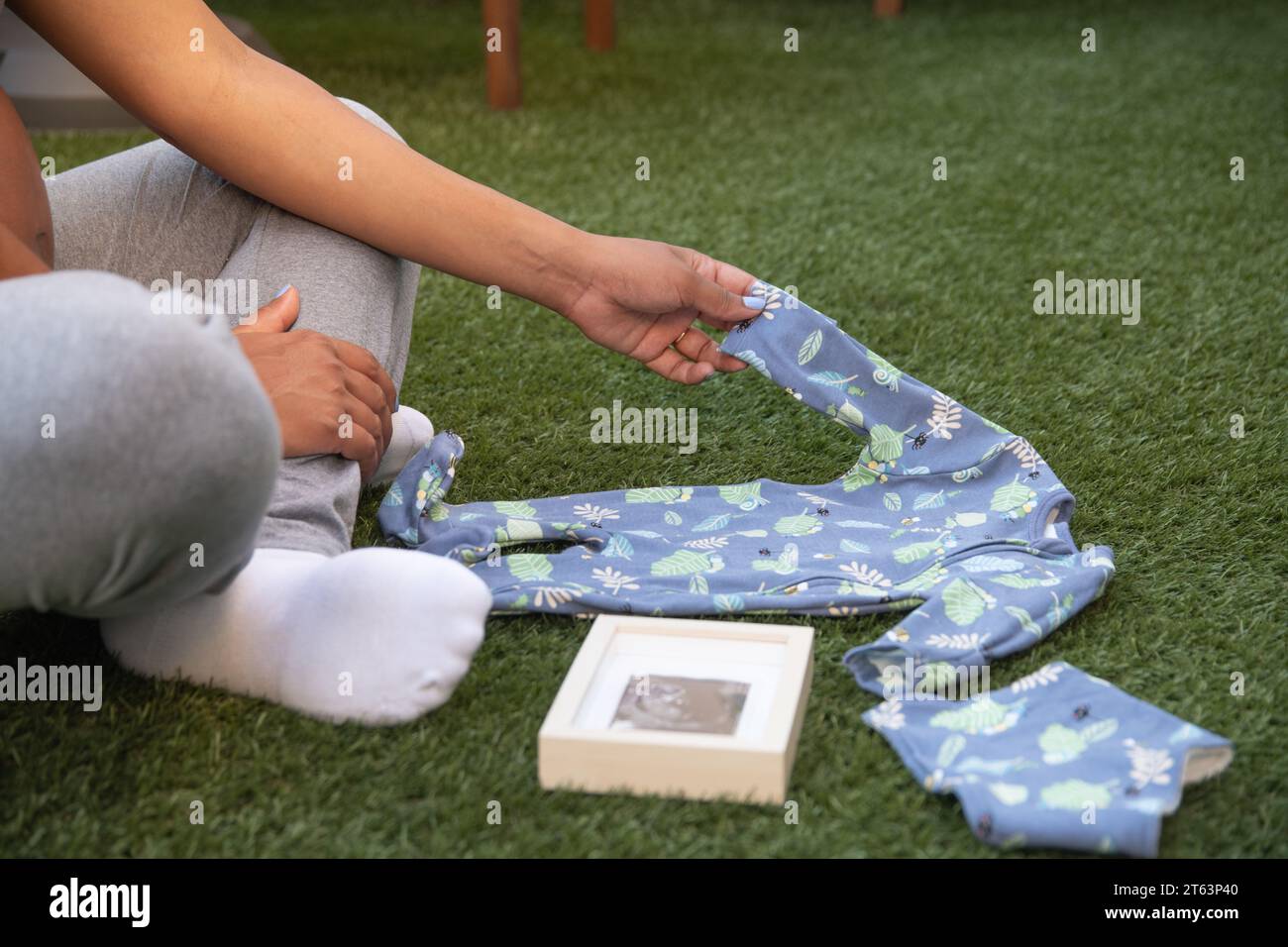 La vista ritagliata di una donna incinta in un sereno giardino organizza una onesie blu con un'immagine sonogramma accanto a lei, raffigurando il suo affetto materno Foto Stock