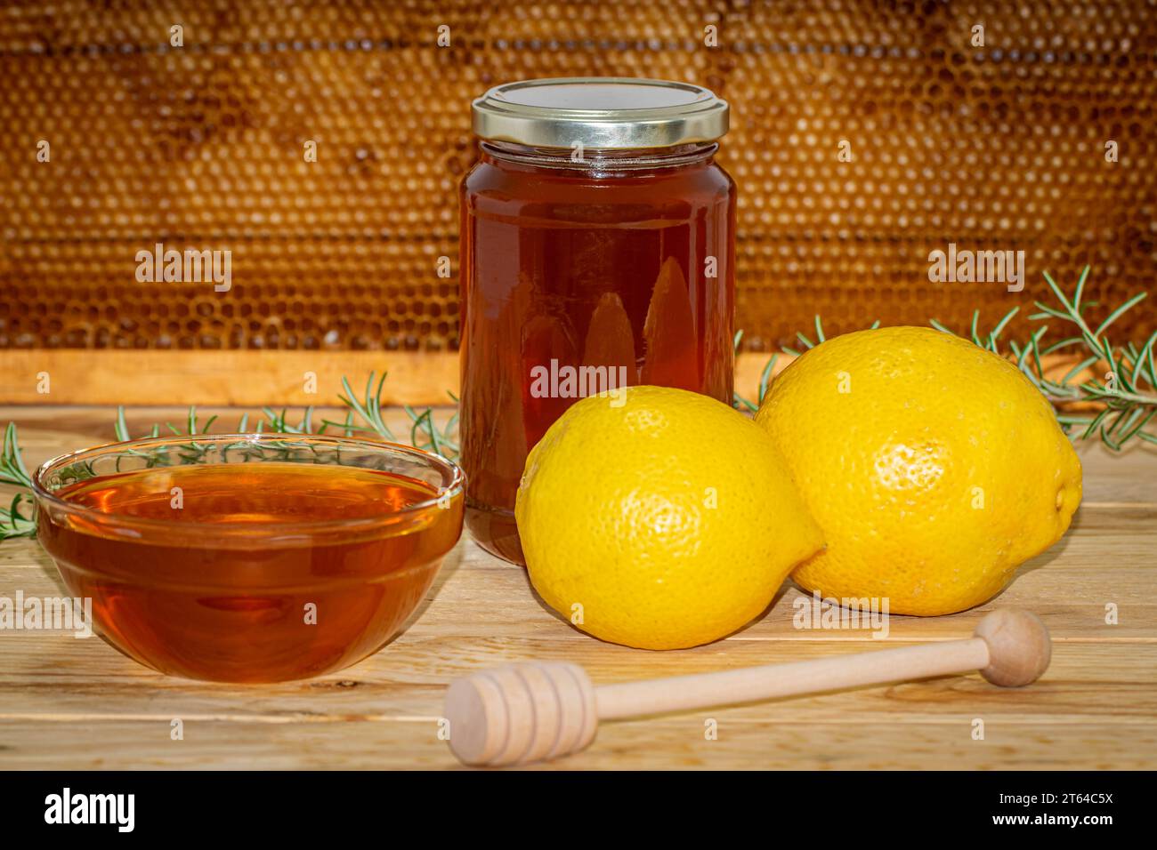 Vaso di miele, dispenser, recipiente con miele, rametto di rosmarino e due limoni su legno con un vero favo sullo sfondo Foto Stock