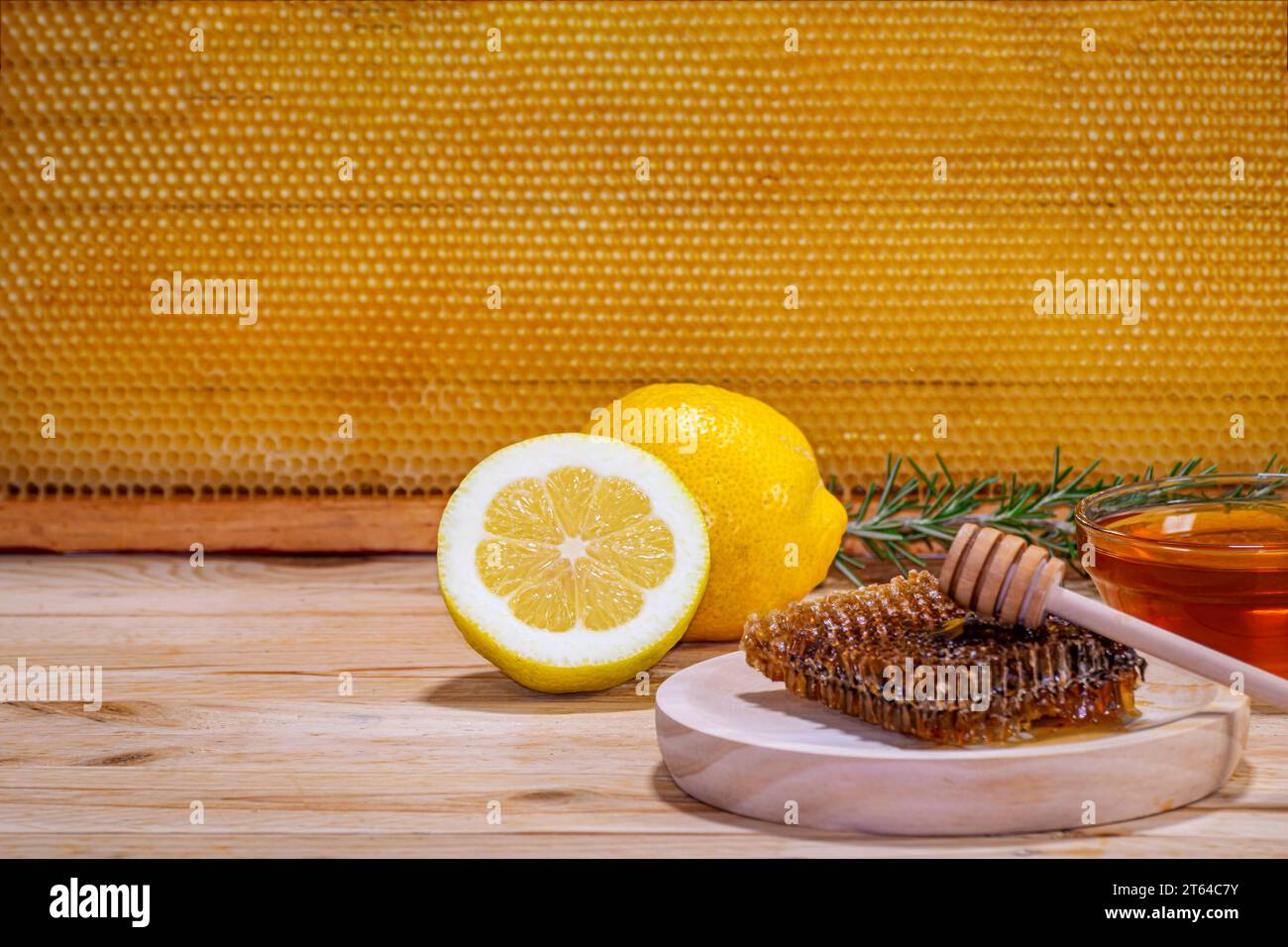 Pezzo di favo su un piatto di legno con un rametto di rosmarino, una ciotola con miele, un limone e mezzo e un favo sullo sfondo. Spazio per il testo Foto Stock