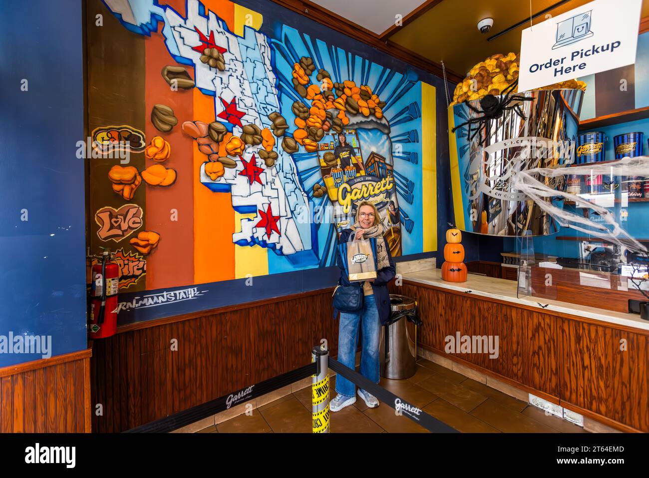 La giornalista Angela Berg si trova nel negozio principale di Garrett Popcorn su Michigan Avenue a Chicago, Illinois, Stati Uniti, davanti a un murale con motivi di Chicago, tenendo un sacchetto di carta con il logo della catena di popcorn, fondata nel 1949. A destra sono visibili decorazioni per Halloween, tra cui piccole lanterne jack-o’-lantern, una ragnatela e un ragno di plastica. Foto Stock