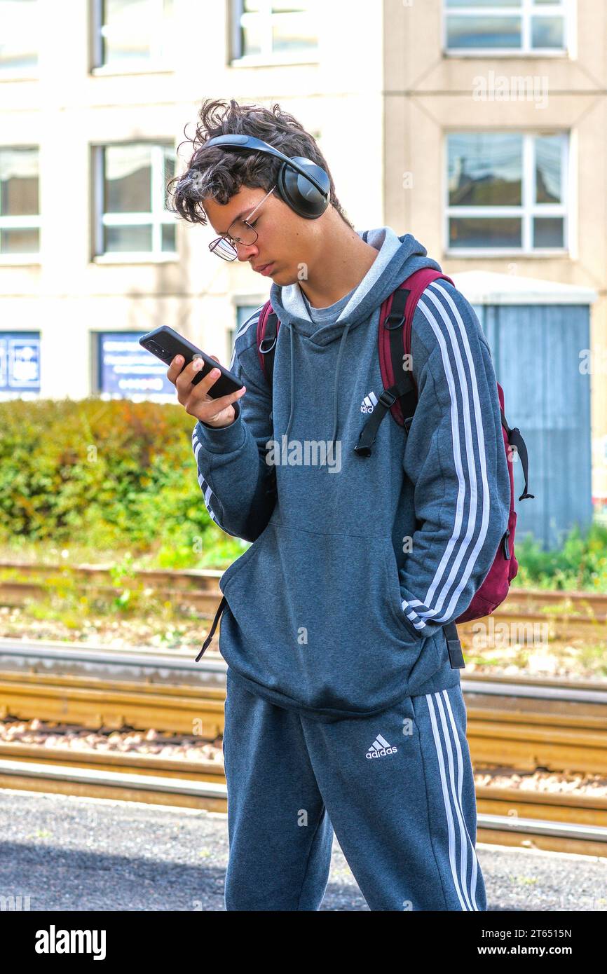 Giovane uomo che indossa abbigliamento sportivo Adidas con smartphone e cuffie BOSE in attesa di treno - Tours, Indre-et-Loire (37), Francia. Foto Stock