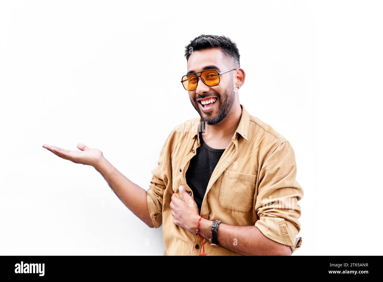 Giovane uomo latino sorridente allegro che presenta e punta con il palmo della mano guardando la fotocamera. Pubblicità, posizionamento dei prodotti e mano su bac bianco Foto Stock