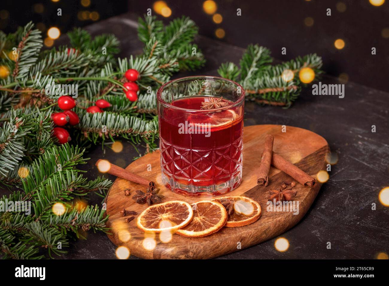 VIN brulé in un bicchiere su tagliere di legno con spezie, rami di abete rosso e luci bokeh, primo piano. Foto Stock