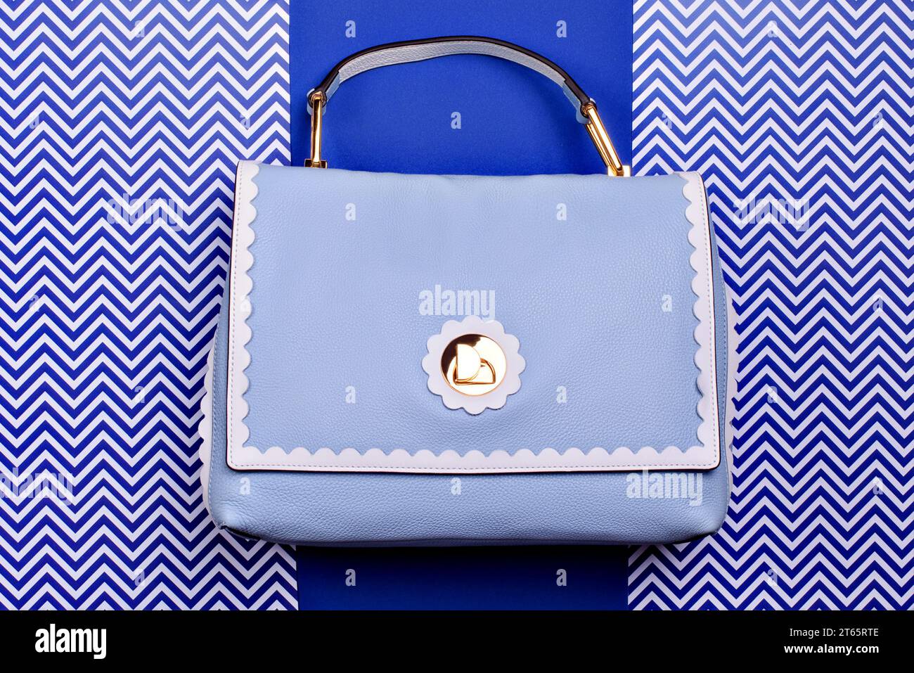 Elegante borsa da donna con manico superiore in pelle blu con dettagli decorativi ondulati e dettagli in metallo dorato su sfondo blu creativo con motivo a zig-zag bianco. Foto Stock