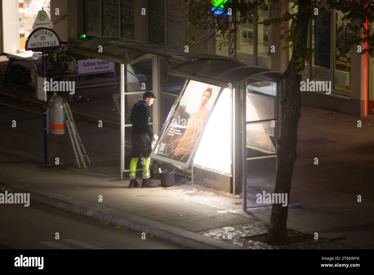 Un cameriere in abiti neri con stivali gialli dotato di secchio sta pulendo una fermata dell'autobus di notte in inverno, Vienna, Austria Foto Stock