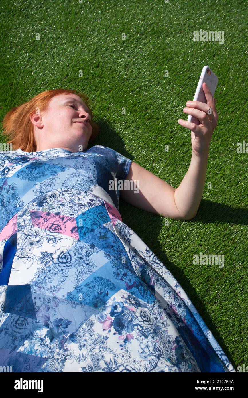 Una donna dai capelli rossi che indossa un abito blu a motivi geometrici, sdraiata a faccia in su e sorridente sull'erba artificiale al sole, guardando il suo telefono Foto Stock