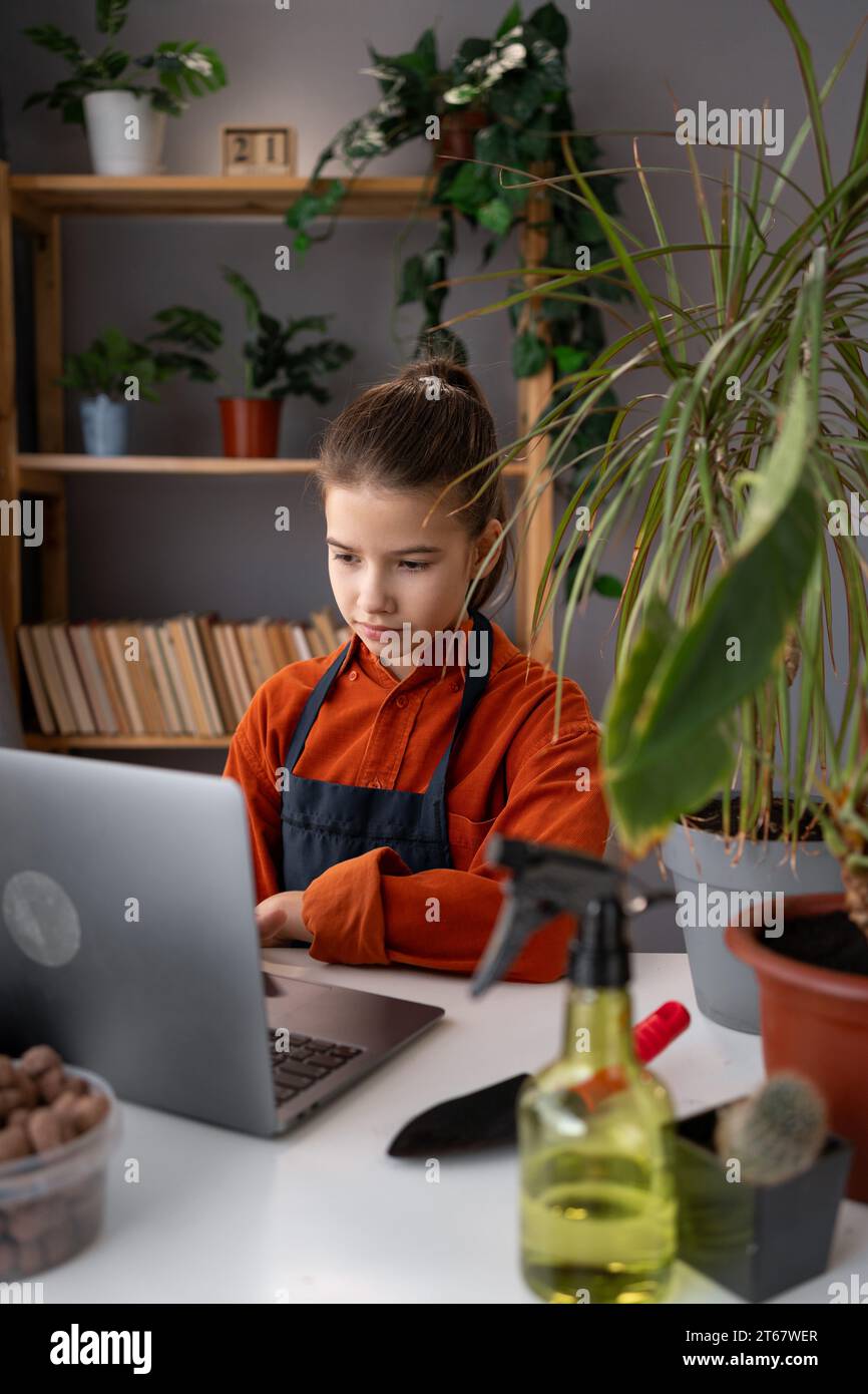 piantagione primaverile: una ragazza adolescente trapianta fiori al coperto a casa mentre esplora un nuovo hobby con il suo laptop. apprendimento pratico e verde Foto Stock