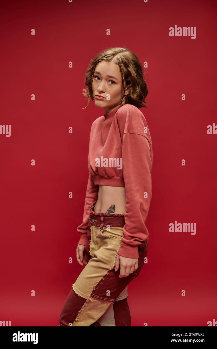 look innocente, giovane donna con pantaloni patchwork e maniche lunghe tagliate in posa su sfondo rosso Foto Stock