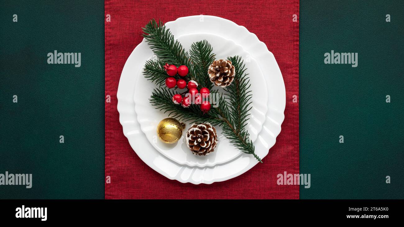 Tavolo di Natale con decorazioni, piatto bianco vuoto su tovaglia rossa, sfondo verde, vista dall'alto, sopra. Festeggiamenti natalizi: Allestimento artistico, rosso A. Foto Stock