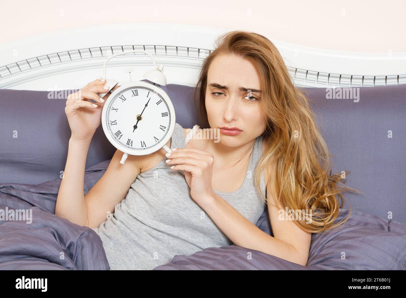 Una giovane donna scioccata che si sveglia con l'allarme, ha dormito troppo, ha dormito male insonnia. La ragazza tiene l'orologio quando è sdraiata a letto la mattina Foto Stock