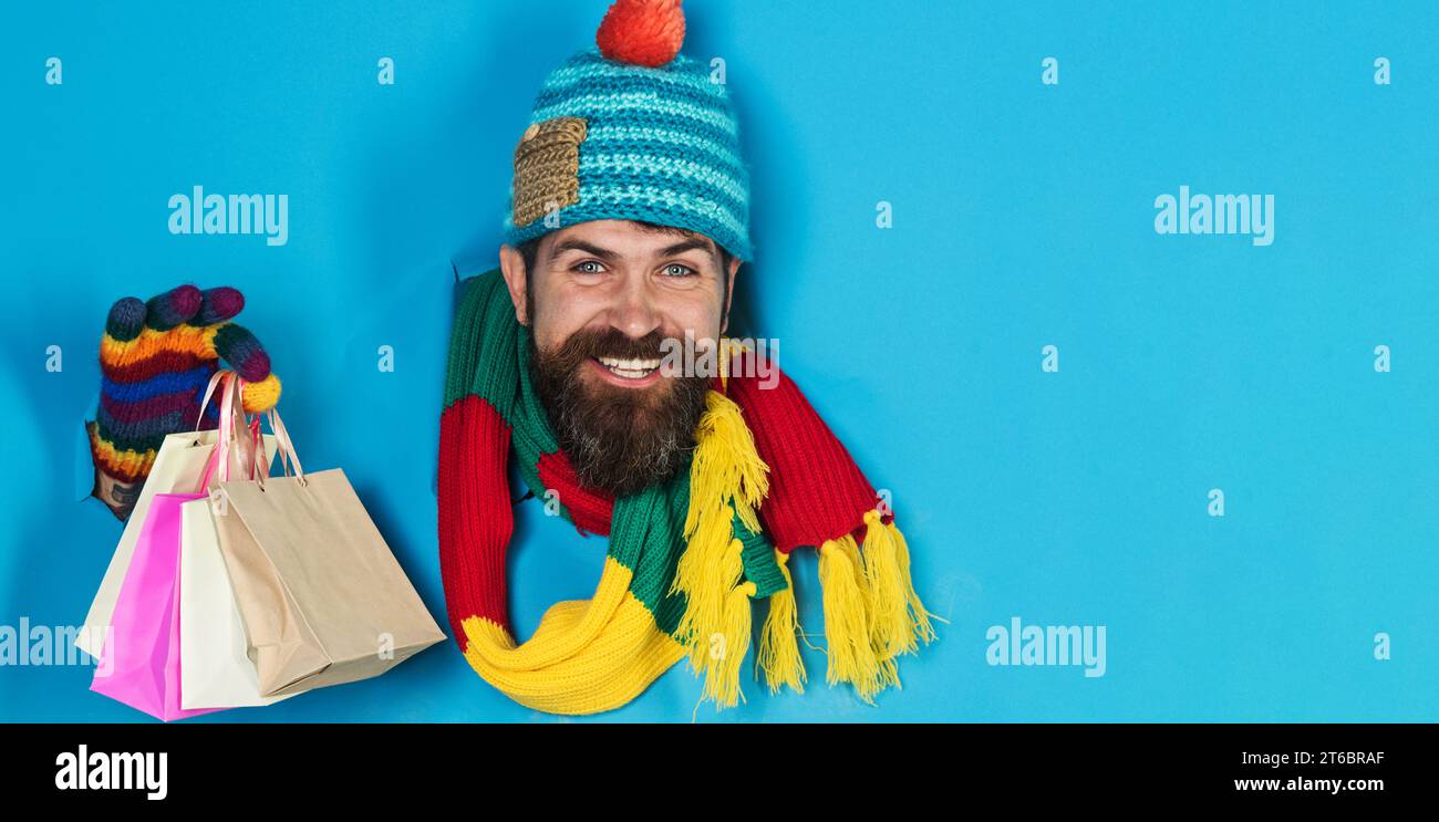Uomo barbato sorridente in abiti invernali con borse della spesa che guardano attraverso il buco della carta. Bel maschio in cappello colorato, sciarpa e guanti con piccolo regalo Foto Stock