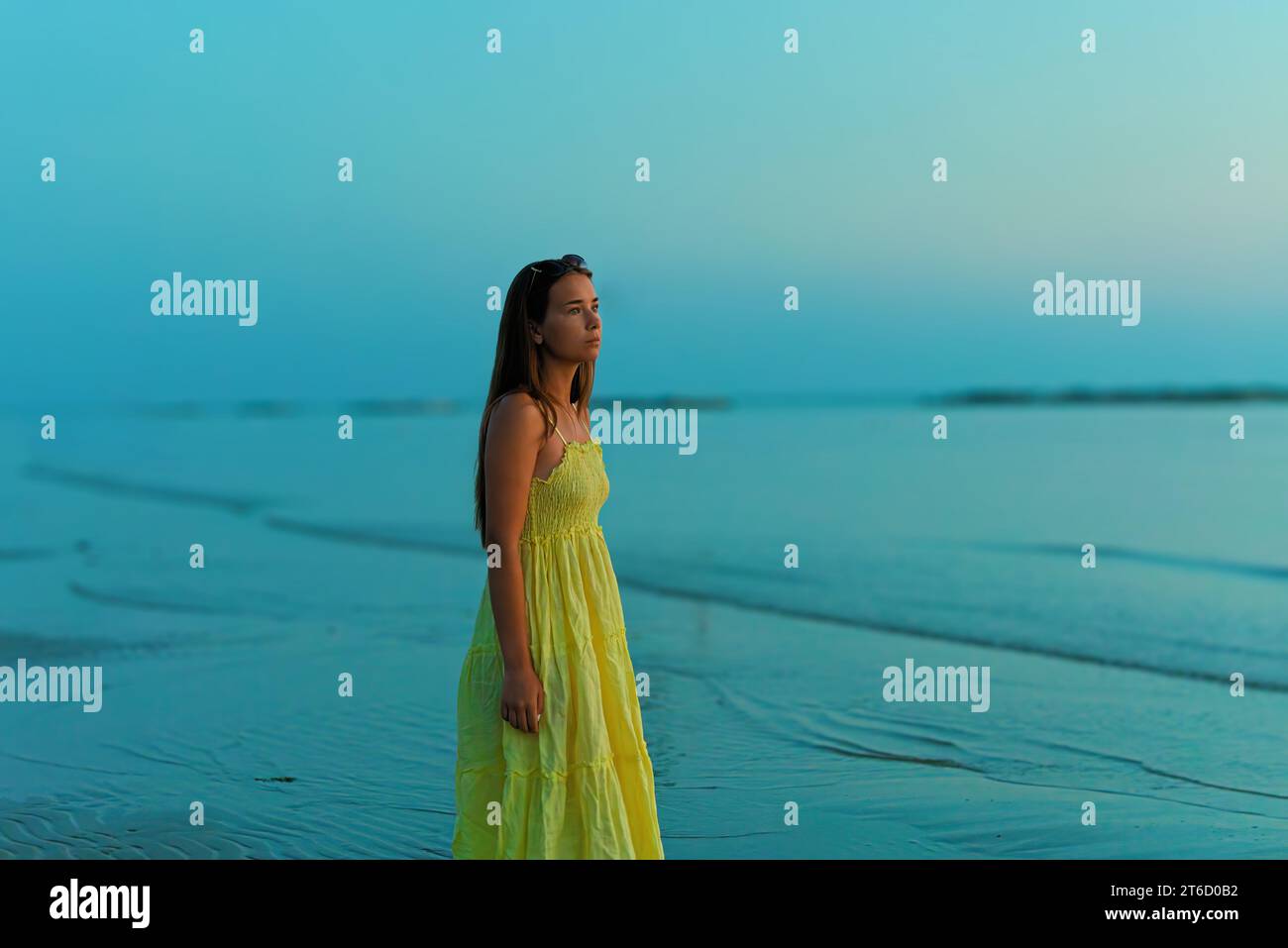 Una giovane donna con un vestito giallo, che guarda con ansia l'alba, cammina lungo la spiaggia. Foto Stock