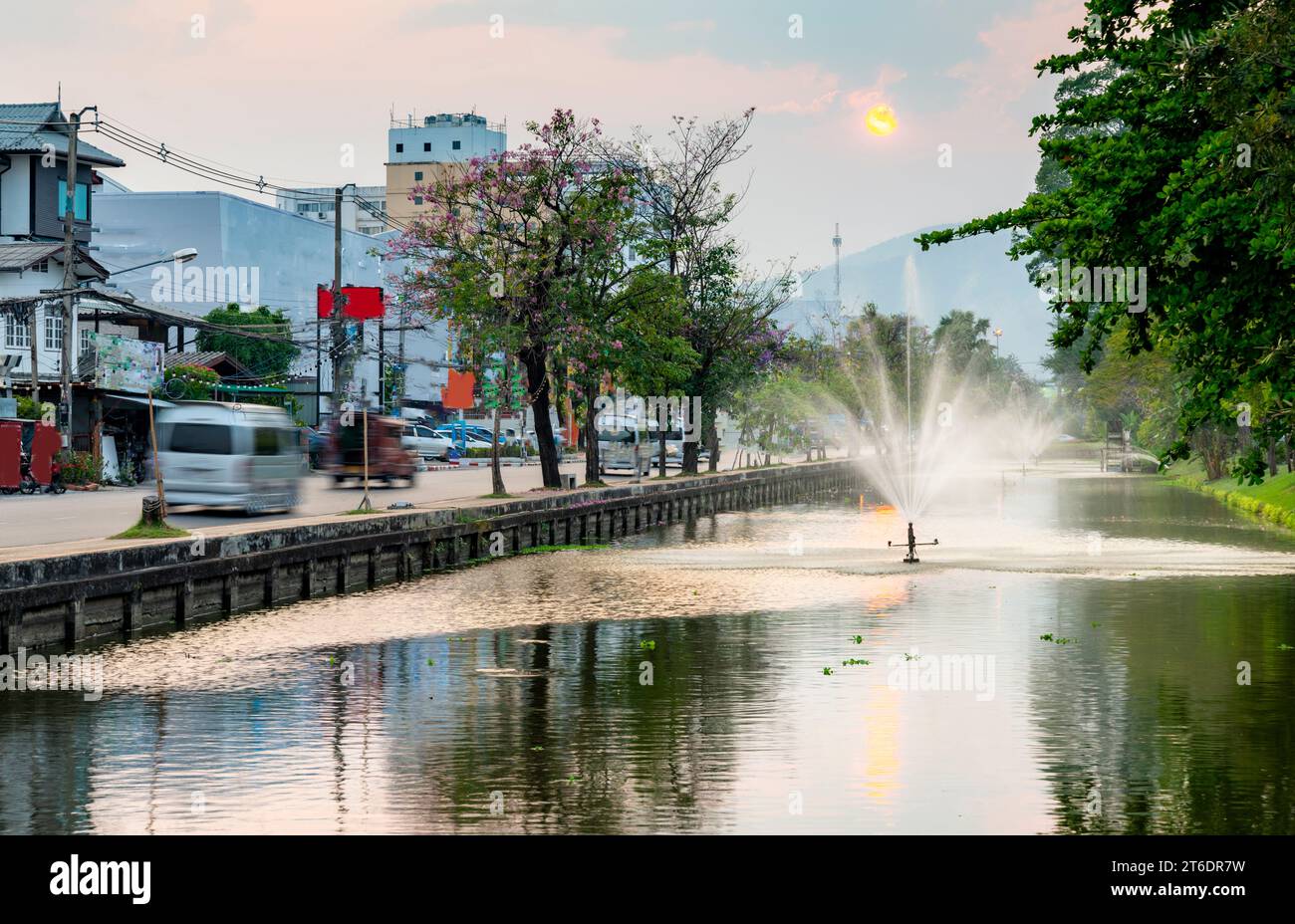 Gli Srinklers spruzzano acqua sul corpo d'acqua che circonda il perimetro della città Vecchia della capitale settentrionale della Thailandia. Il sole tramonta su montagne affumicate Foto Stock