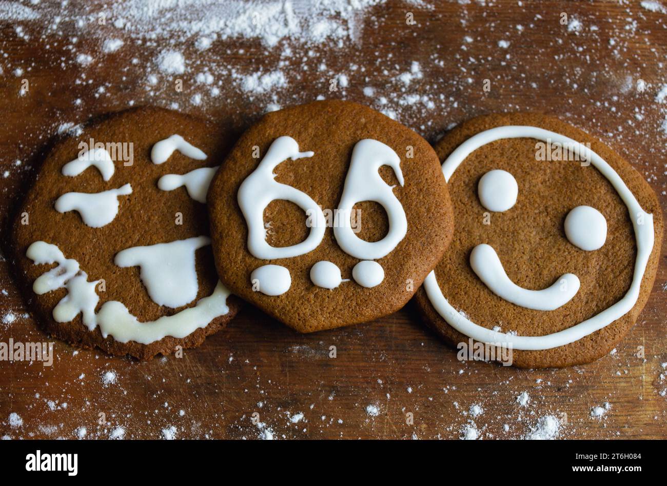 Tre biscotti dolci ricoperti di glassa bianca sotto forma di emoticon e il numero 66 giacciono su una vecchia tavola circondata da farina Foto Stock