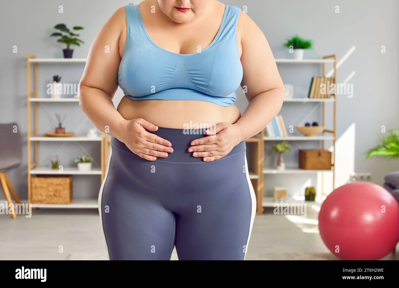 Donna grassa e voluminosa che indossa abbigliamento sportivo guarda purtroppo la sua grande vita in piedi a casa. Foto Stock