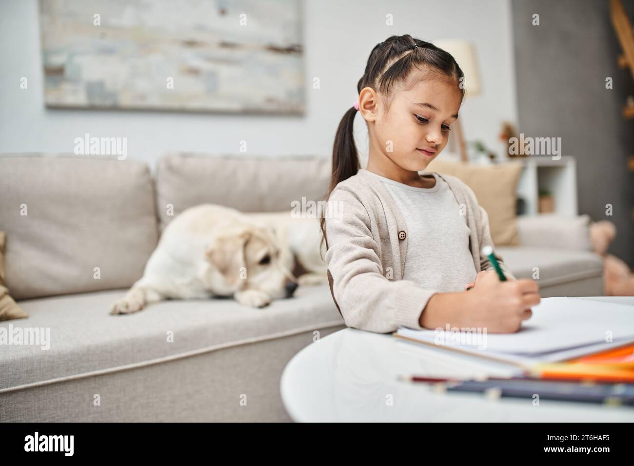 bambina in abiti informali che disegnano su carta con matita colorata vicino al labrador in salotto, arte Foto Stock