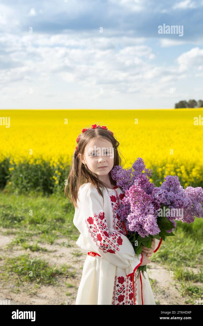 Bella ragazza di 6-7 anni in abiti nazionali ucraini con un bouquet di lilacs sullo sfondo di un fiorente campo di colza gialla. Ucraina Foto Stock