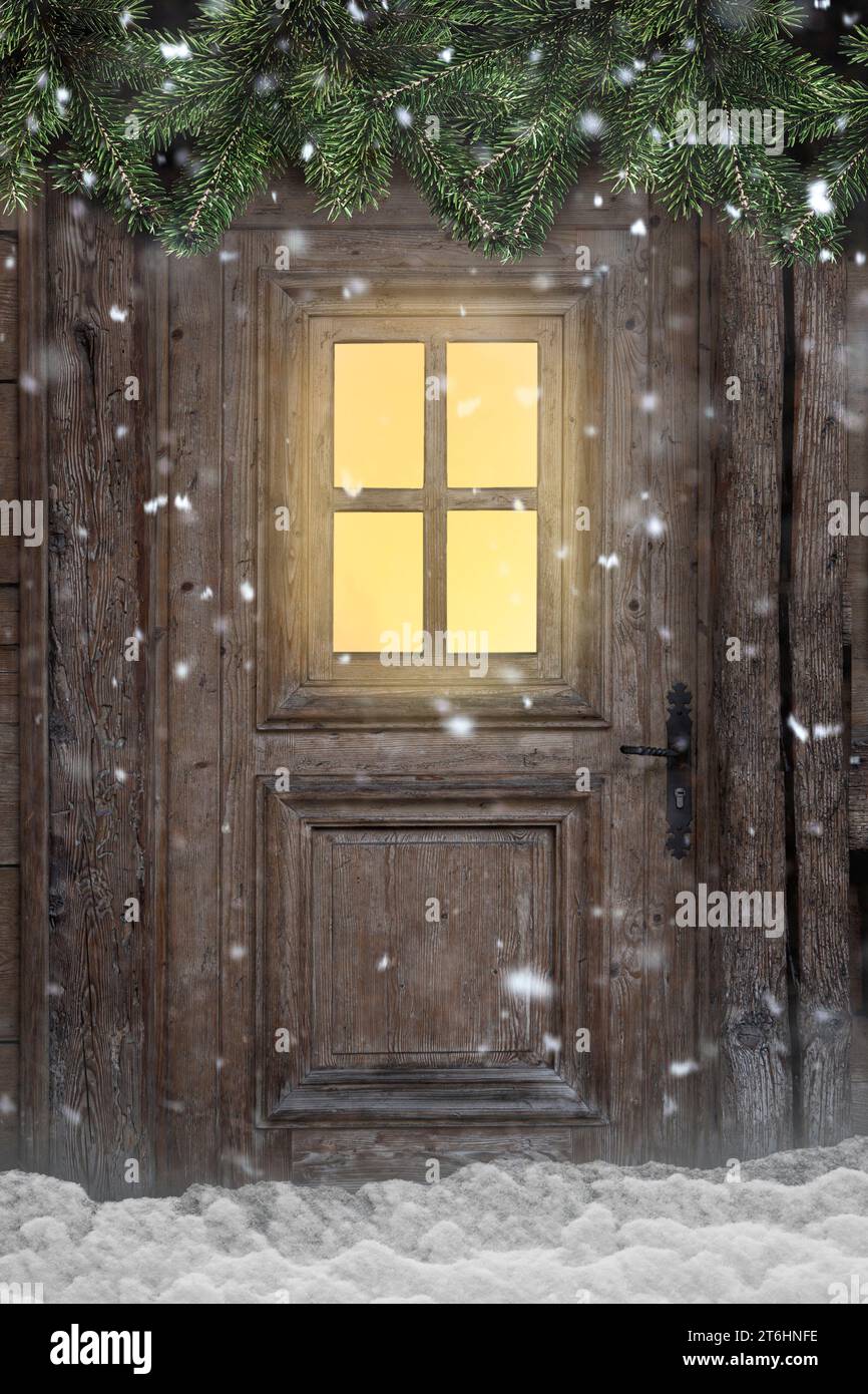 Porta con finestra illuminata in un rifugio innevato con rami di abete Foto Stock