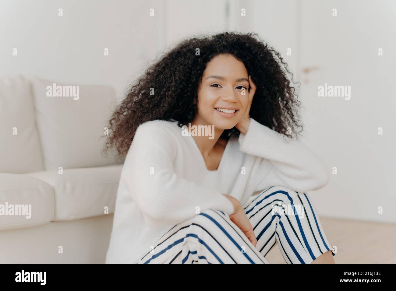 Donna sorridente con voluminosi capelli ricci seduta in uno spazio luminoso, vestita con abiti comodi Foto Stock