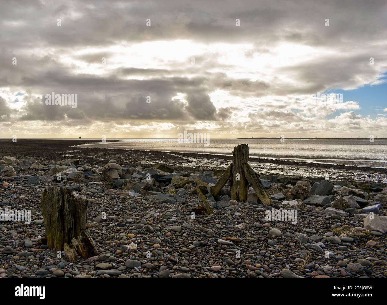 Una spiaggia di rocce sciolte e ciottoli con i resti di una struttura in legno, in lontananza c'è una bassa marea sotto un cielo nuvoloso ma luminoso. Foto Stock