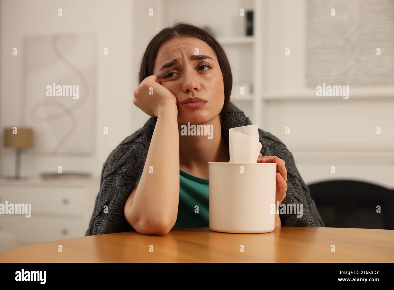 Donna malata avvolta in coperta con scatola di fazzoletti al tavolo di legno al chiuso. Sintomi del raffreddore Foto Stock