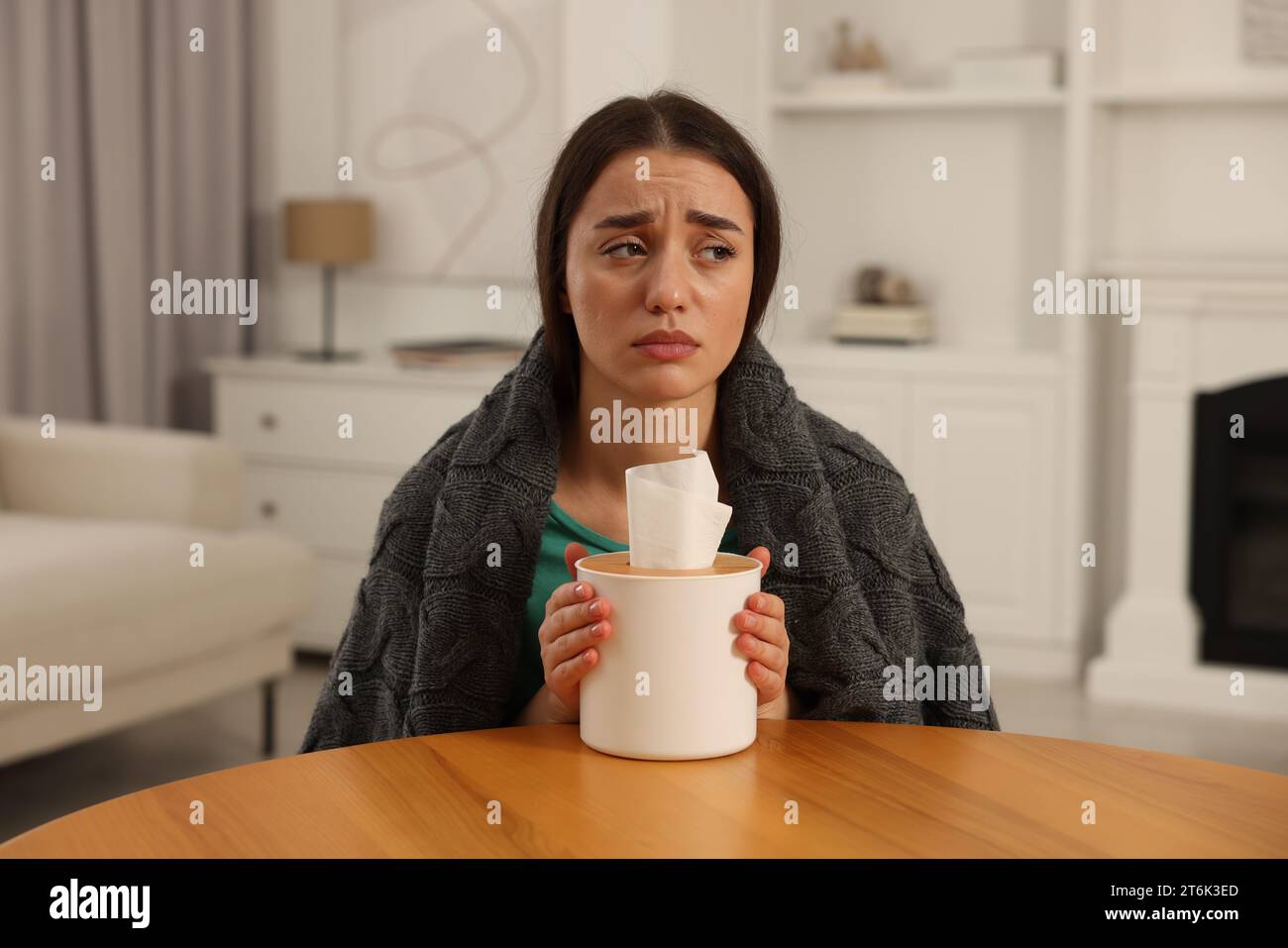 Donna malata avvolta in coperta con scatola di fazzoletti al tavolo di legno al chiuso. Sintomi del raffreddore Foto Stock