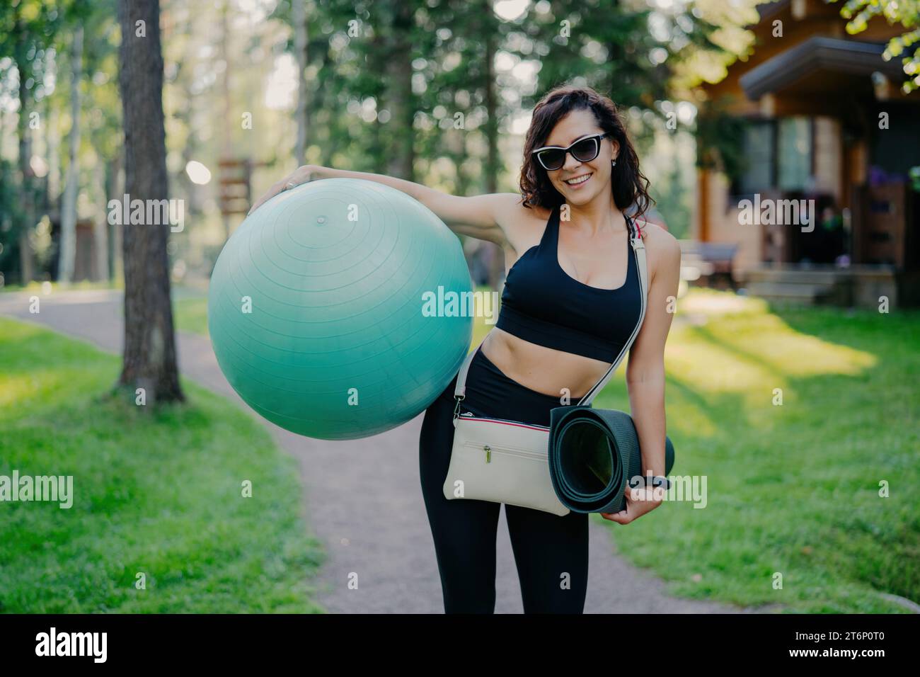 Donna attiva e radiosa con palla da ginnastica e tappetino yoga, con un sorriso soleggiato in un parco lussureggiante Foto Stock