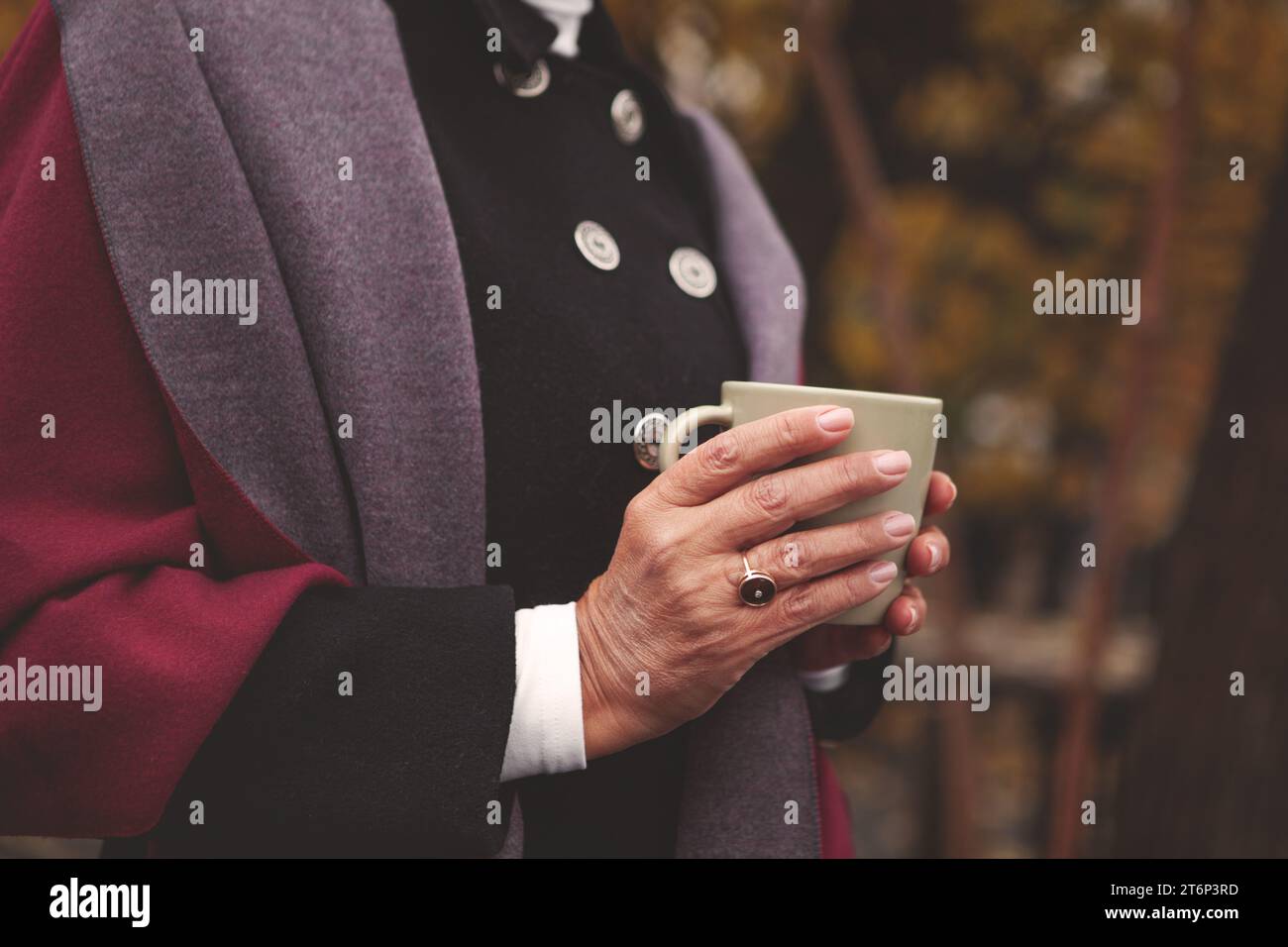 Mani ravvicinate di una donna anziana, vestita comodamente, che tiene una tazza con caffè o tè sullo sfondo della natura autunnale. Atmosfera rilassante, atmosfera calda Foto Stock