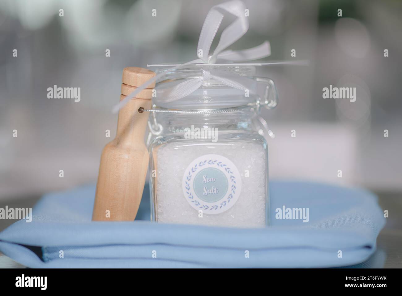 Bellissimo vaso trasparente riempito di sale marino con fiocco bianco e legna su coperta blu davanti al bokeh bianco Foto Stock