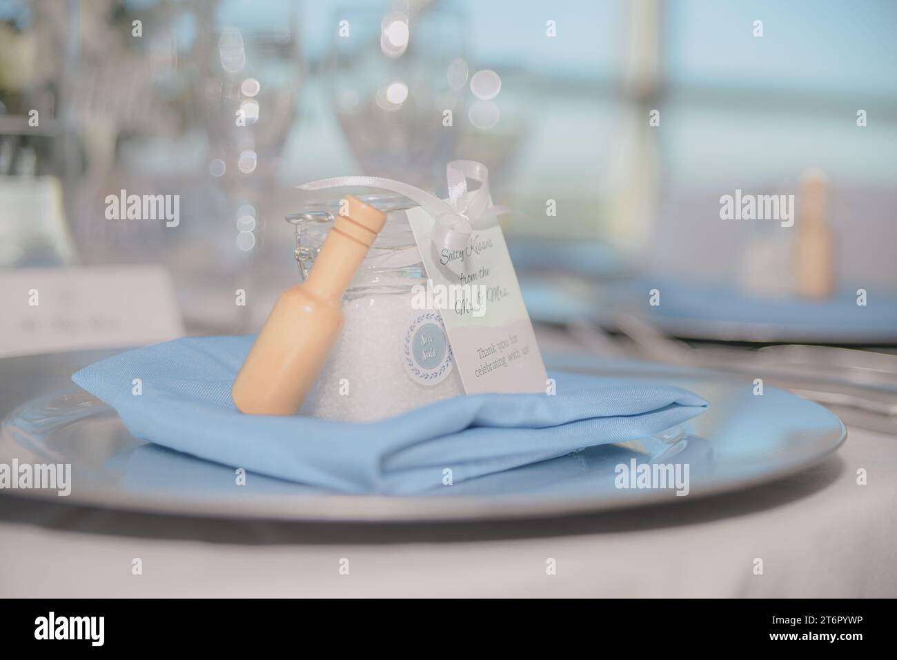 Bellissimo vaso trasparente riempito di sale marino con fiocco bianco e legna su coperta blu su trey di fronte al bokeh bianco Foto Stock