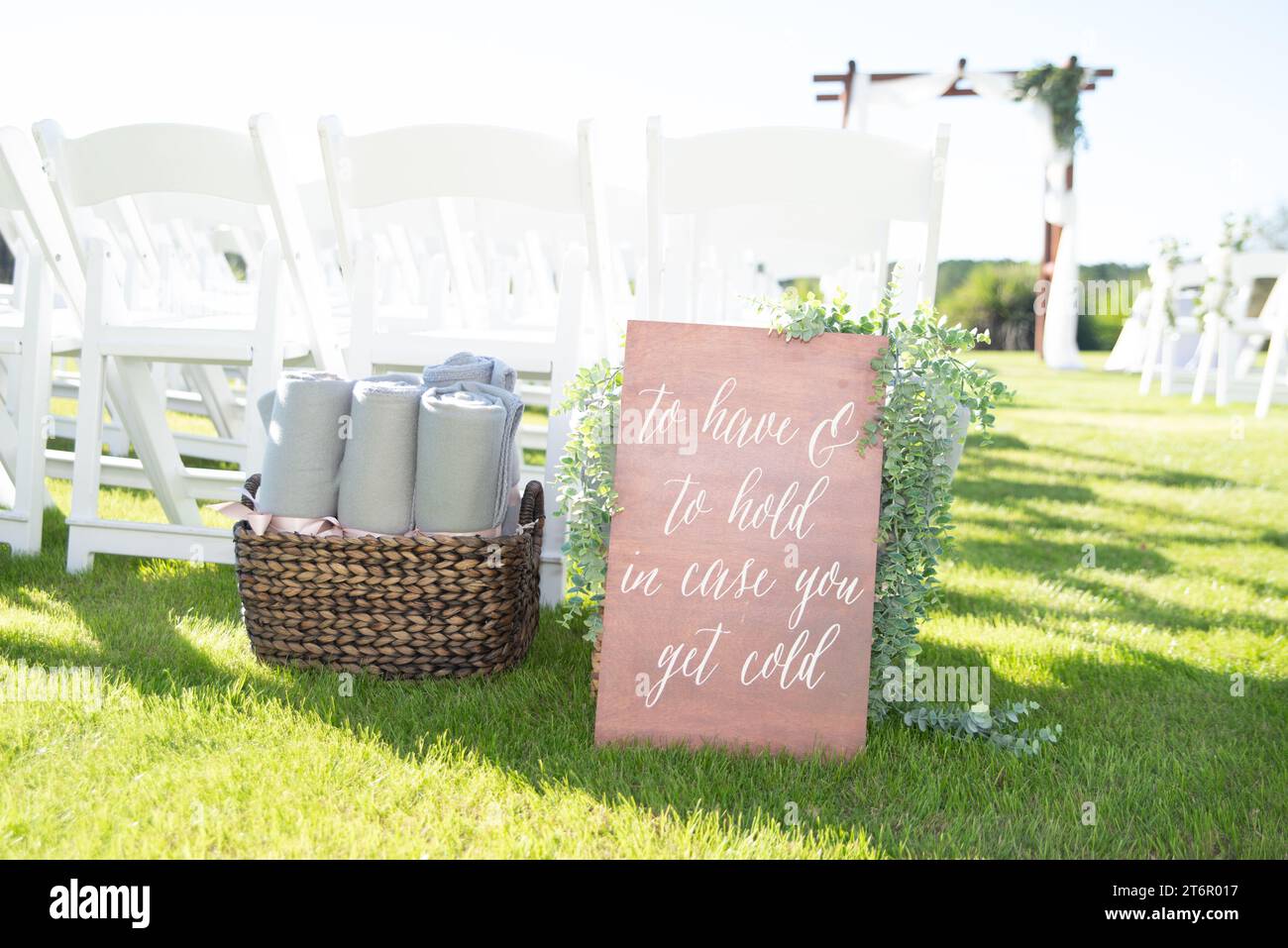 Un cartello di legno che recita "da avere e da custodire, si fa freddo" sull'erba durante la cerimonia di nozze davanti alle sedie bianche accanto al cestino di vimini con asciugamani Foto Stock