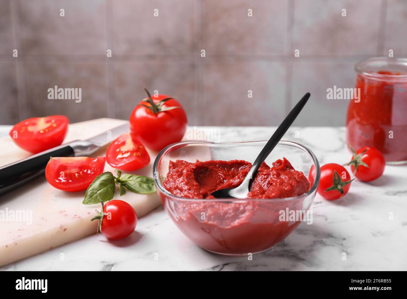 Ciotola in vetro di gustosa pasta di pomodoro con cucchiaio e ingredienti su un tavolo in marmo bianco Foto Stock