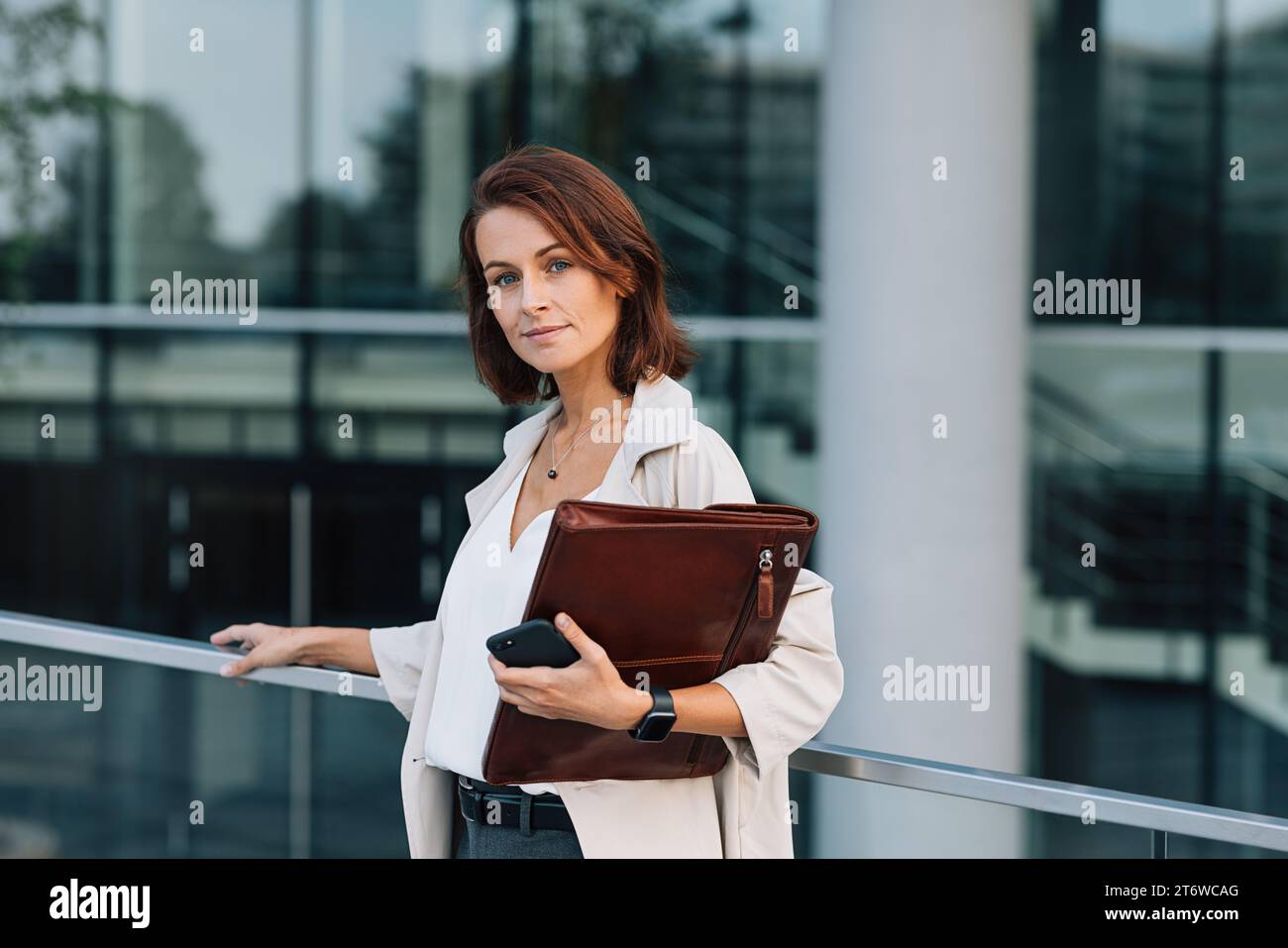 Donna d'affari sicura di sé con capelli di zenzero che guarda la fotocamera. Donna di mezza età con cartella all'edificio aziendale. Foto Stock
