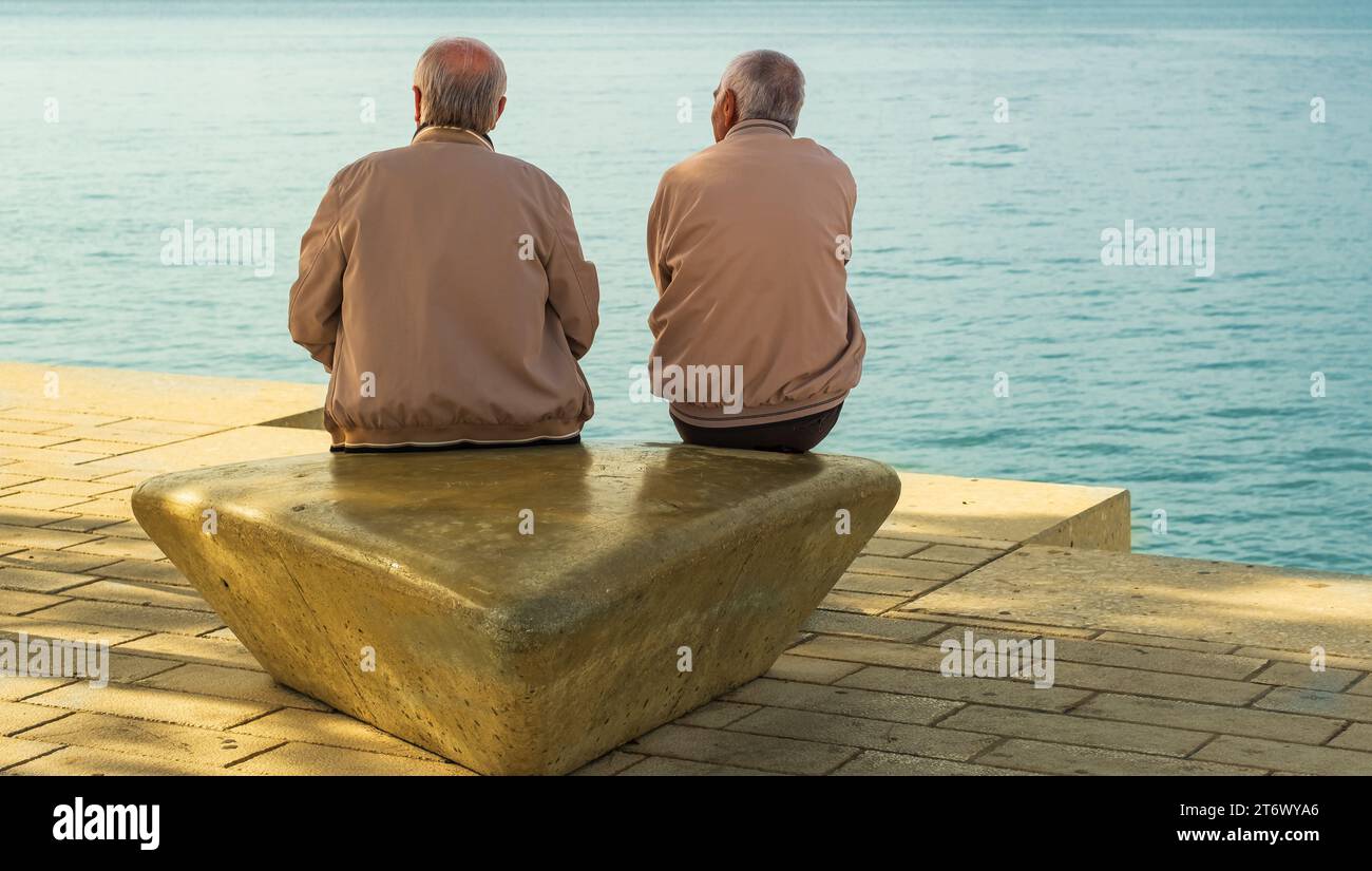 Due amici maschi sulla spiaggia seduti e chiacchierando mentre guardavano l'oceano. Due uomini in pensione seduti su una pietra di marmo, che parlano e si rilassano. Vecchio Foto Stock