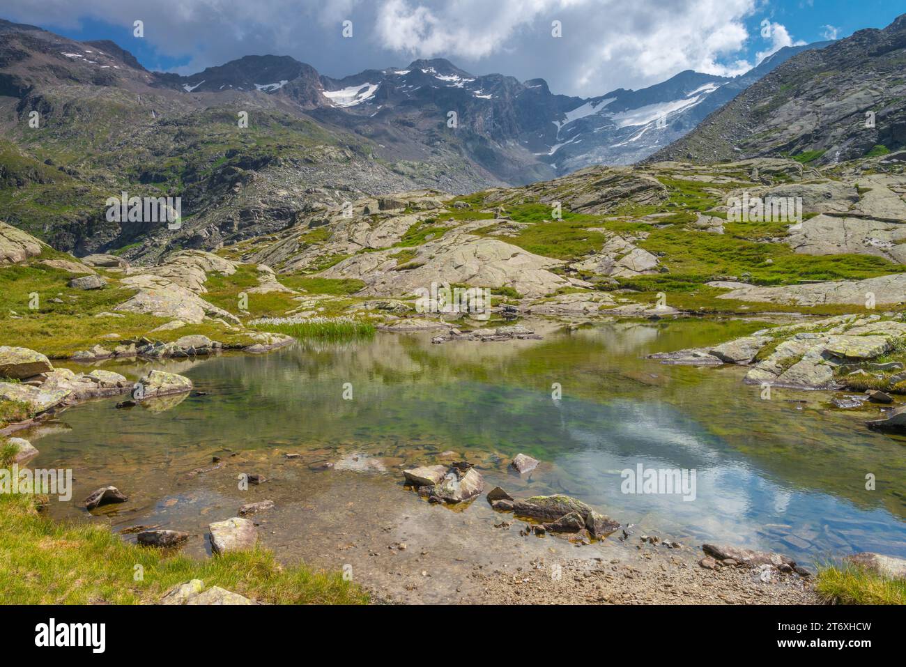 Maestosa scena montuosa nelle Alpi italiane, imponente catena montuosa con un piccolo ghiacciaio che torreggia sulla valle con piccolo stagno alpino e lastre di roccia. Foto Stock