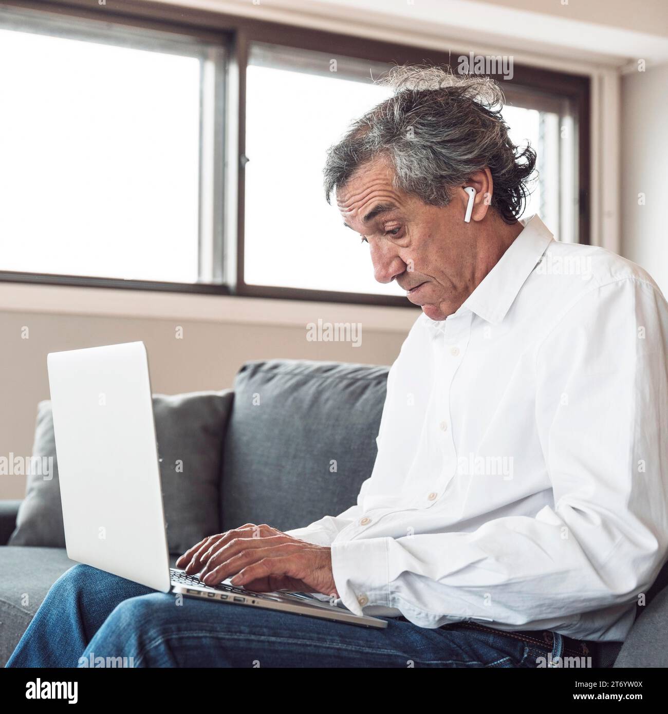 Ritratto uomo anziano seduto divano che indossa auricolari bluetooth con computer portatile Foto Stock