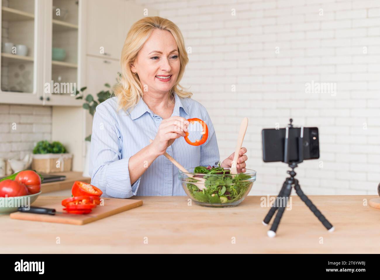 Donna anziana che effettua una videochiamata sul cellulare mostrando una fetta di peperone mentre prepara l'insalata Foto Stock