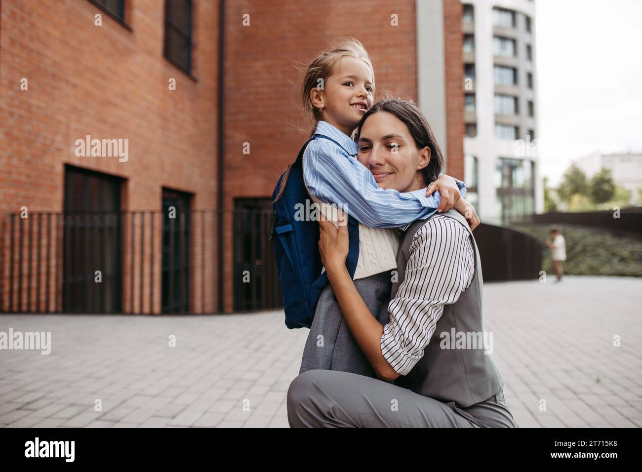 Madre lavoratrice che abbraccia la figlia davanti all'edificio scolastico e si dirige al lavoro. Concetto di equilibrio tra lavoro e vita privata per le donne. Foto Stock